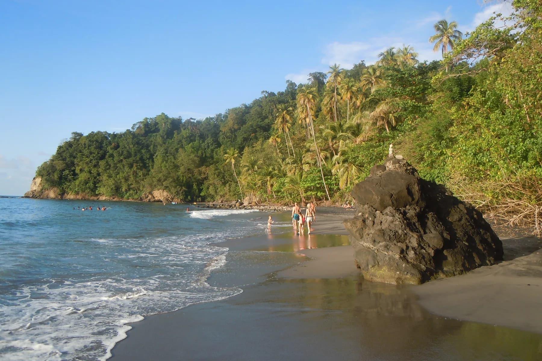 Plage de sable noir de l'Anse Couleuvre au Prêcheur, bordée de forêt tropicale et d'un gros rocher, avec des baigneurs au bord de l'eau