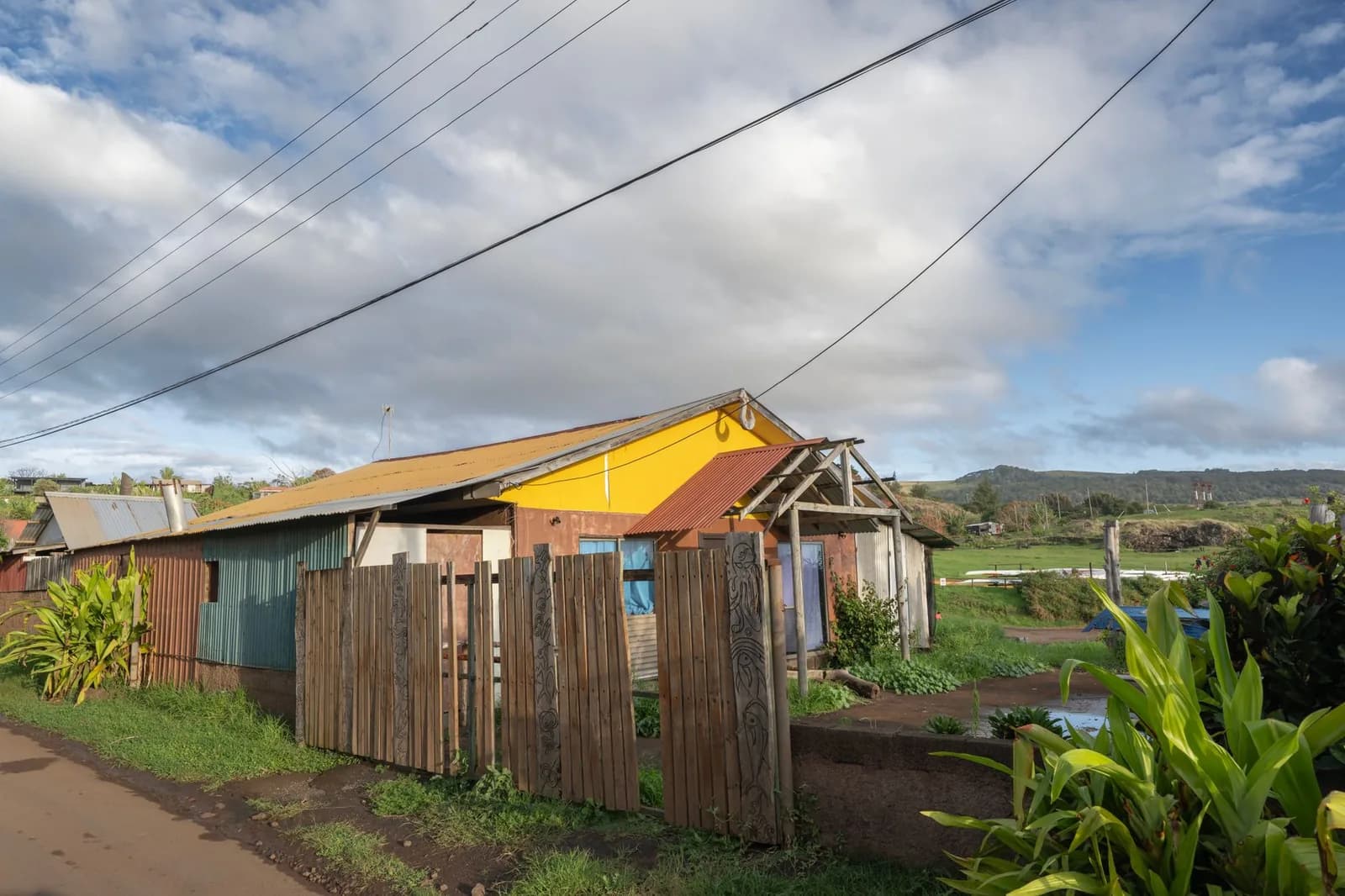 Maison colorée à toit de tôle jaune et rouille derrière une clôture en bois, végétation tropicale et ciel nuageux