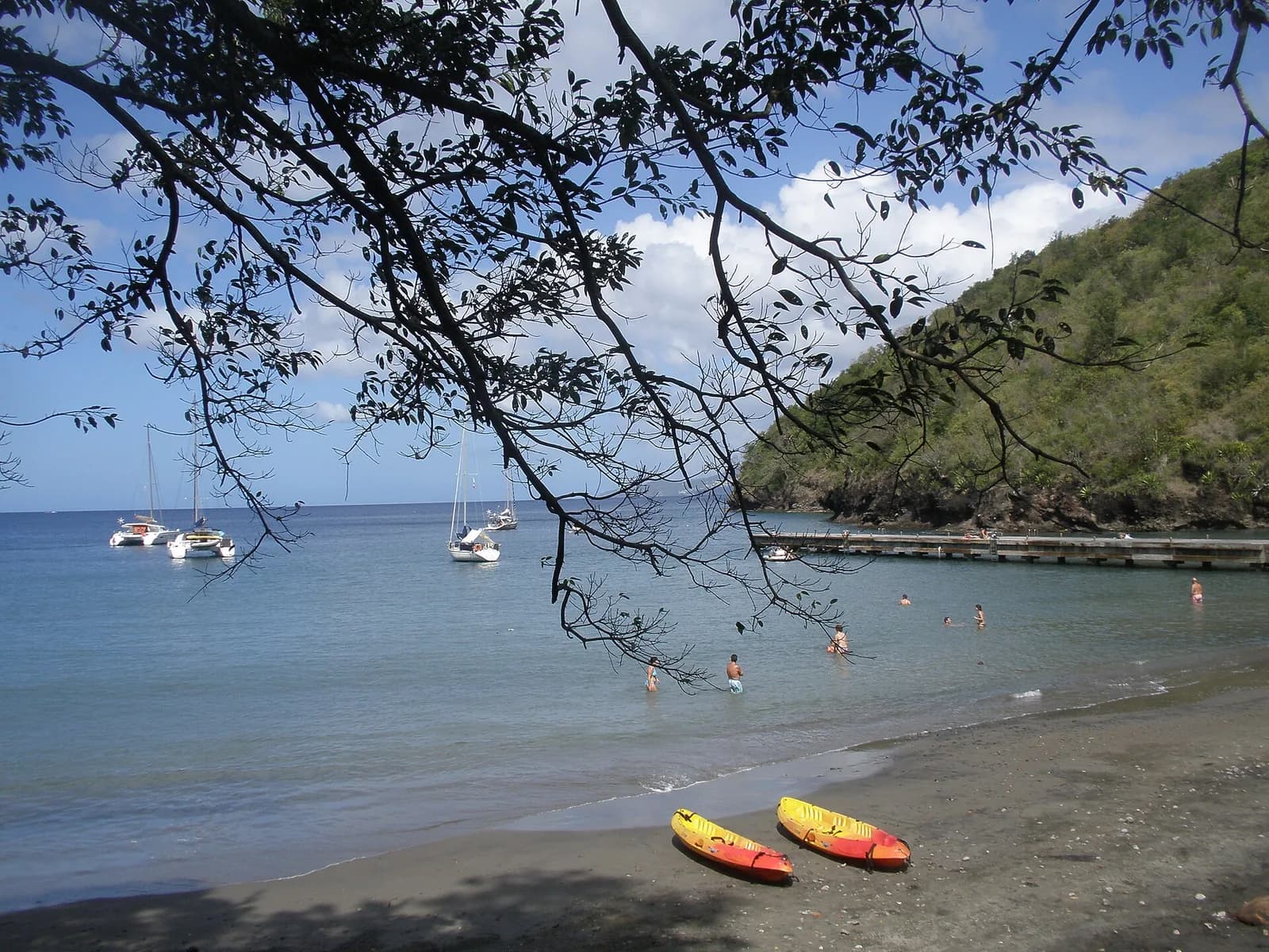 Baie à sable volcanique sombre avec voiliers au mouillage, kayaks sur la plage et colline verte, sur le littoral caraïbe de la Martinique