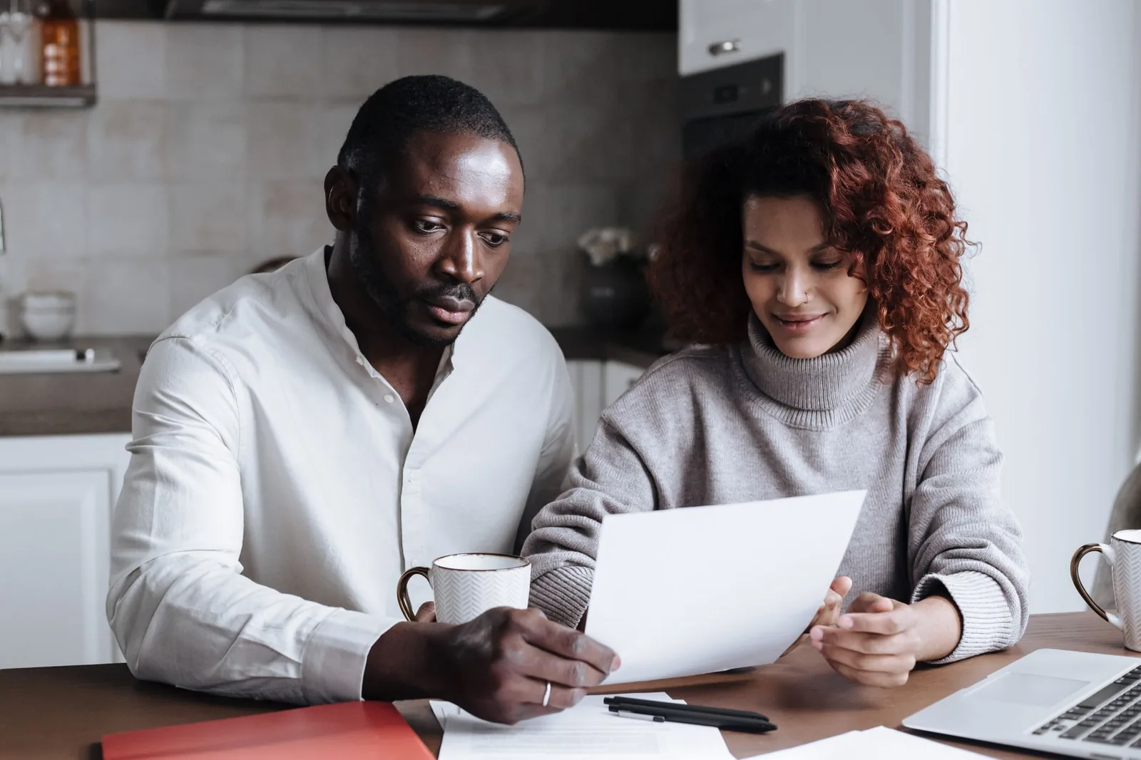 Un couple examine ensemble un document administratif à une table, avec un ordinateur portable et des tasses