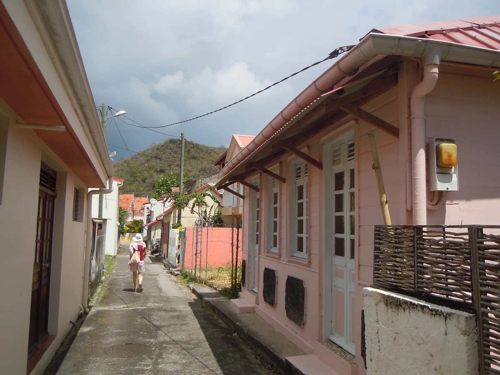 Ruelle bordée de cases créoles aux Anses-d'Arlet, en Martinique, avec un piéton et un morne en arrière-plan