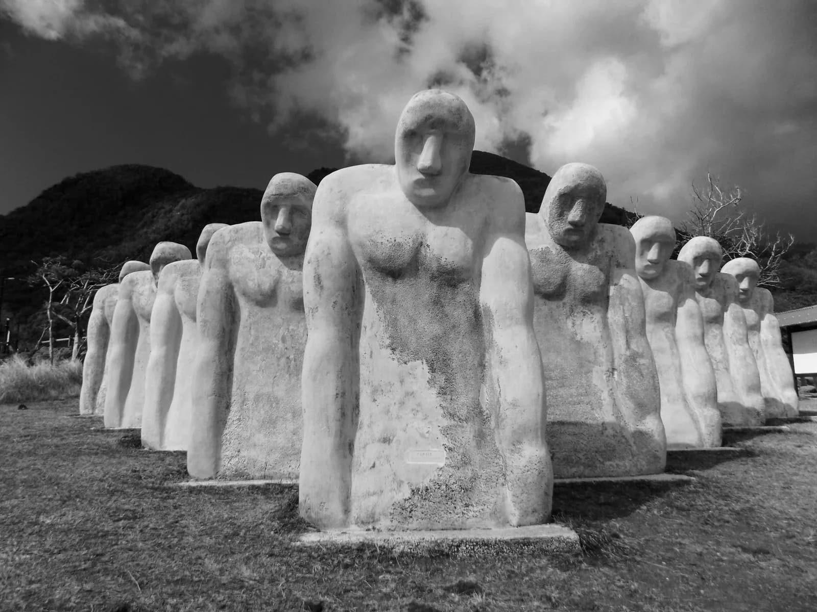 Statues blanches alignées du mémorial Cap 110 devant une colline verte sous un ciel chargé, en noir et blanc