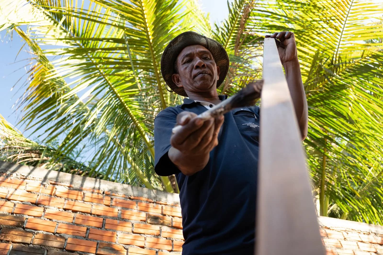 Artisan coiffé d'un chapeau travaillant une planche de bois en extérieur, devant un mur de briques et des palmiers