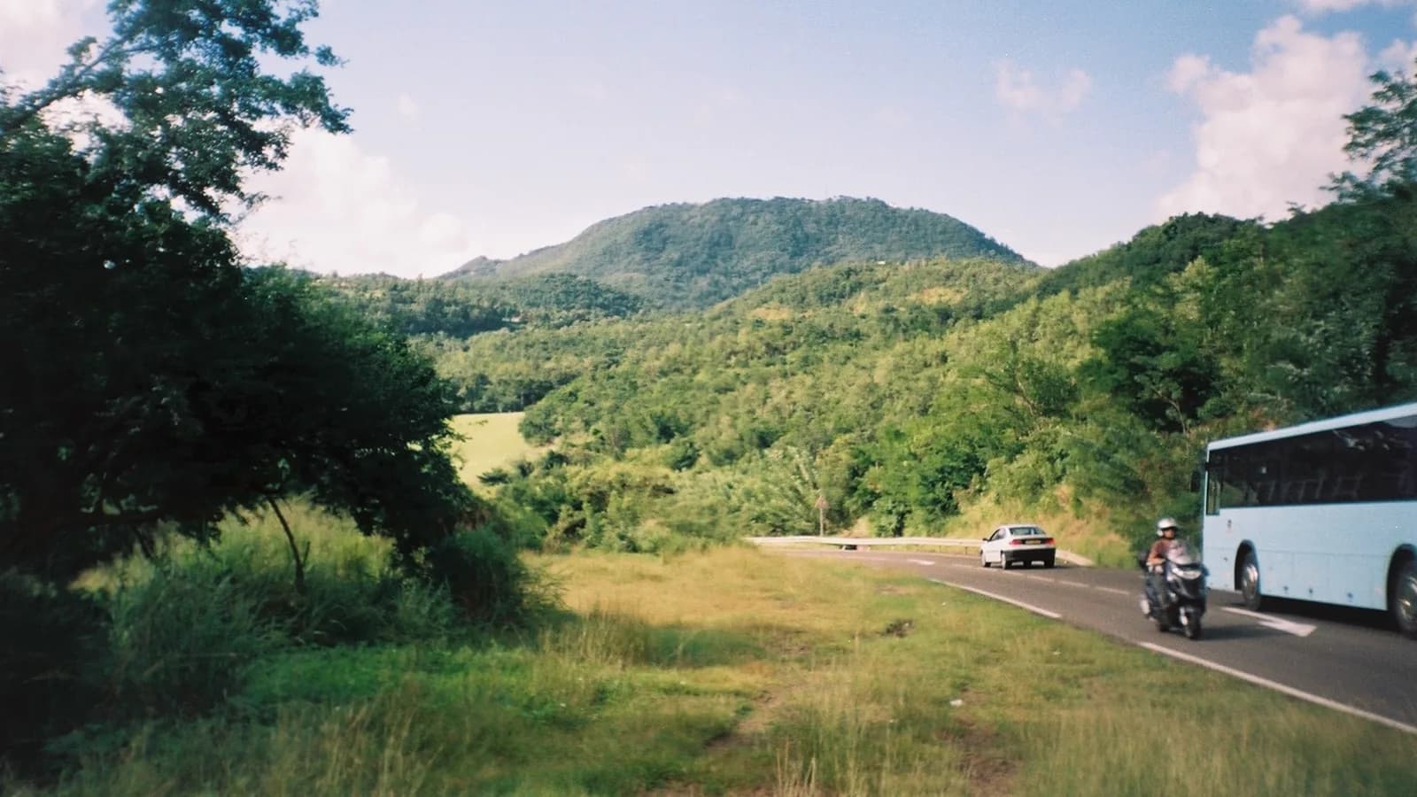 Route départementale de Martinique entre des collines boisées, avec une voiture, un scooter et un car