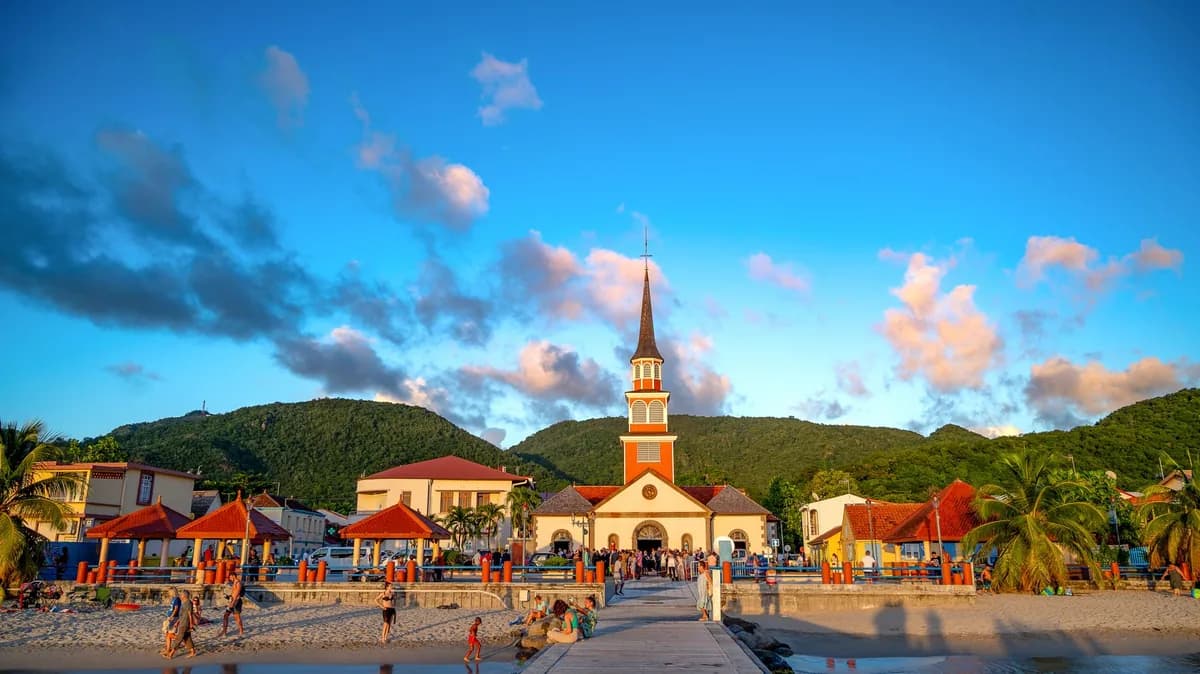 Église au clocher blanc au bord de la plage, maisons colorées et collines vertes aux Anses-d'Arlet