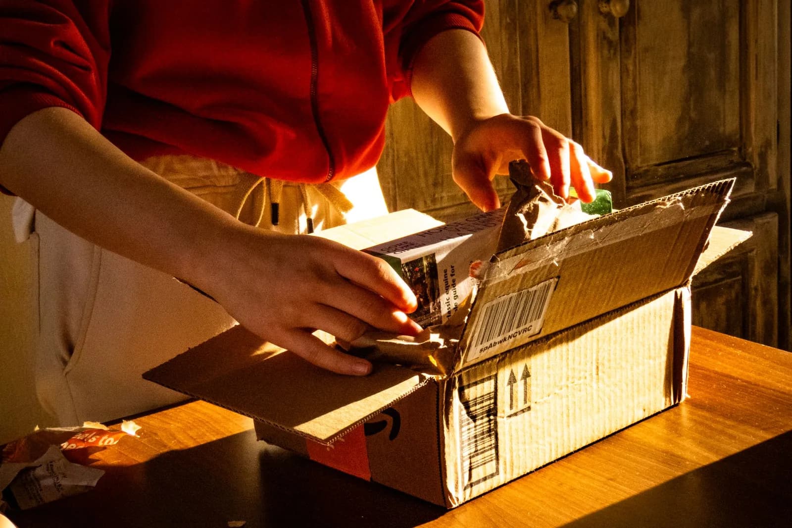 Mains qui ouvrent un colis en carton posé sur une table en bois, à la lumière du soleil