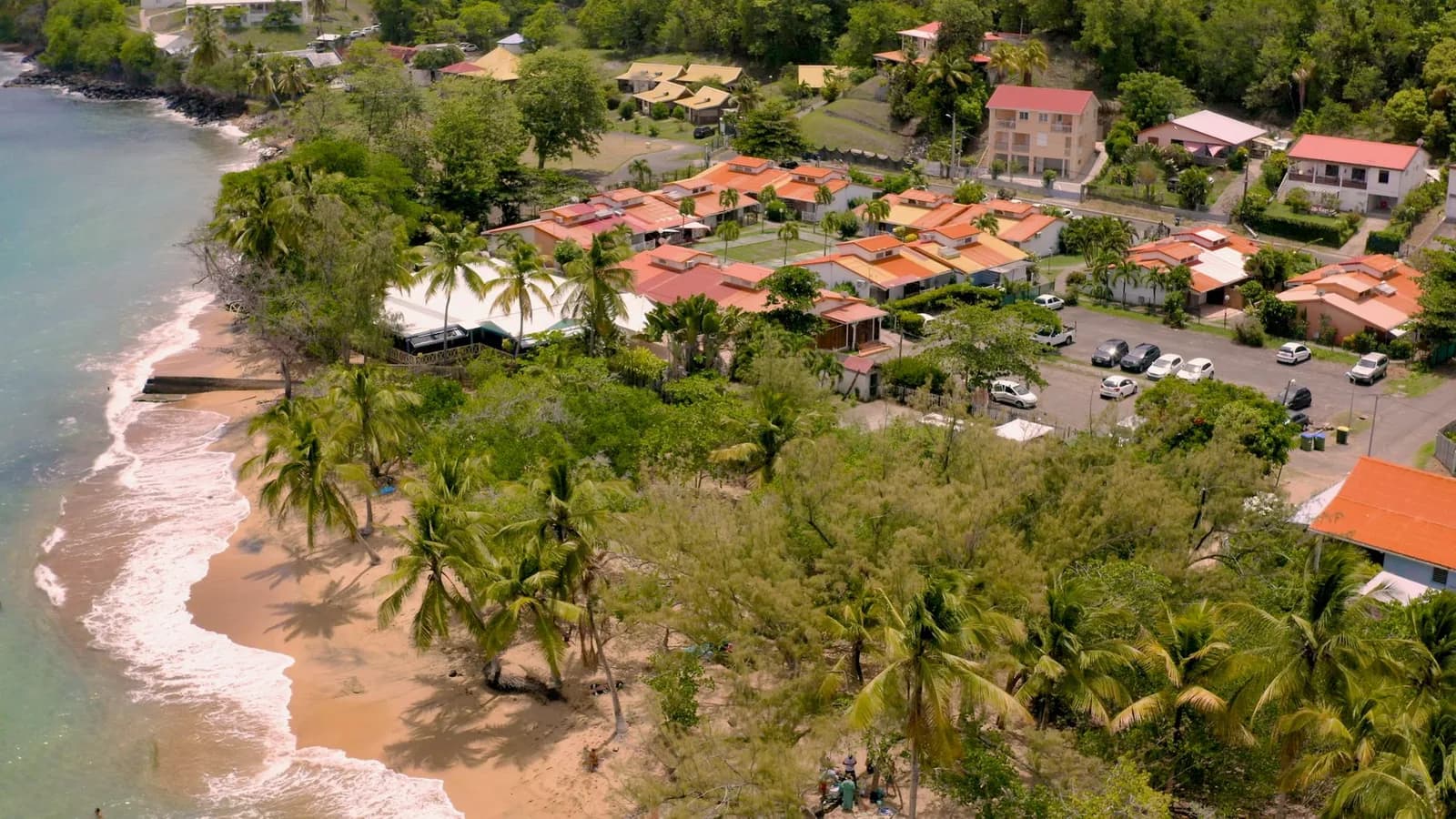 Vue aérienne d'une plage de sable bordée de palmiers et de maisons à toits de tuiles orange, sur la côte sud-caraïbe
