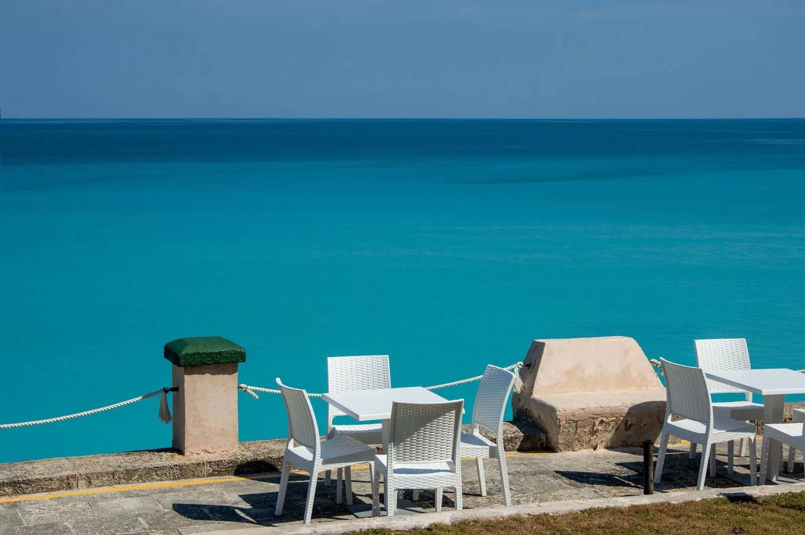 Terrasse de restaurant en bord de mer turquoise, tables et chaises blanches face à l'eau