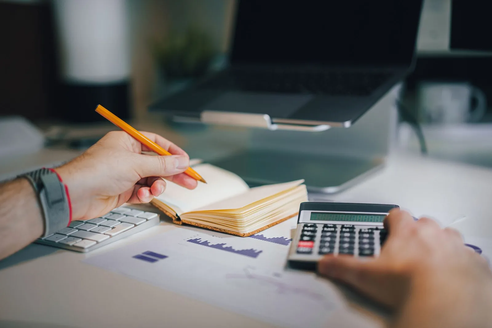 Mains sur un bureau avec une calculatrice, un carnet et des graphiques imprimés pour calculer une rémunération