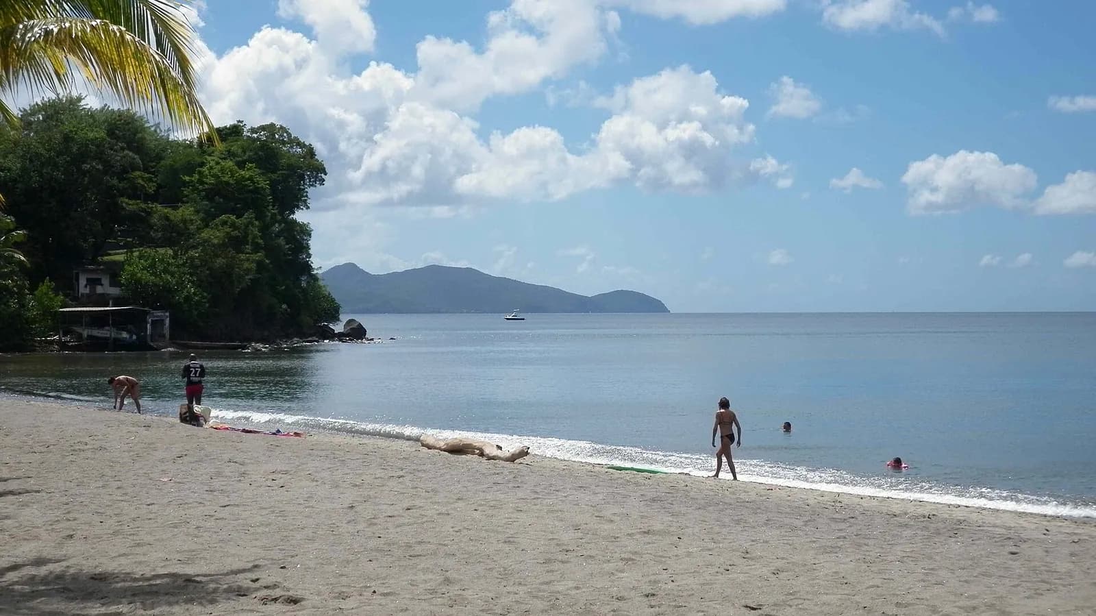 Plage de sable gris de Madiana à Schœlcher, avec des baigneurs, un promontoire boisé et, au loin, les reliefs de la côte caraïbe